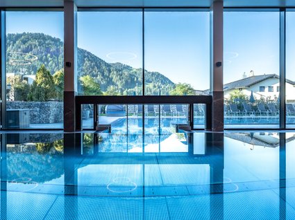 Innenpool mit Bergblick Großer, klarer Innenpool mit Blick durch bodentiefe Fenster auf die umliegende Berglandschaft unter blauem Himmel.