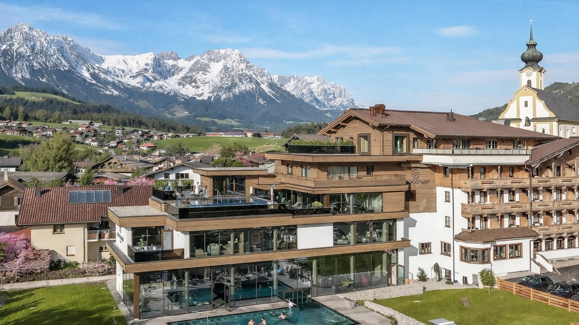 Hotel in Söll am Wilden Kaiser: der Postwirt Modernes Hotel mit Pool vor schneebedeckten Bergen und Dorf im Tal