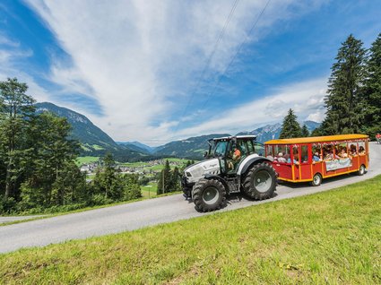 Discover the benefits of the Wilder Kaiser GästeCard to be used in your holiday area- Tractor pulling a red tourist carriage with passengers on a mountain road