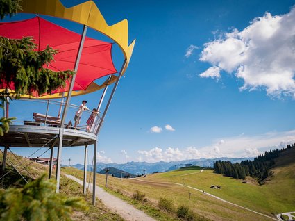 Söll: Ausflugsziele in der Umgebung Menschen auf Aussichtsplattform mit rotem Sonnenschutz und Berglandschaft im Hintergrund