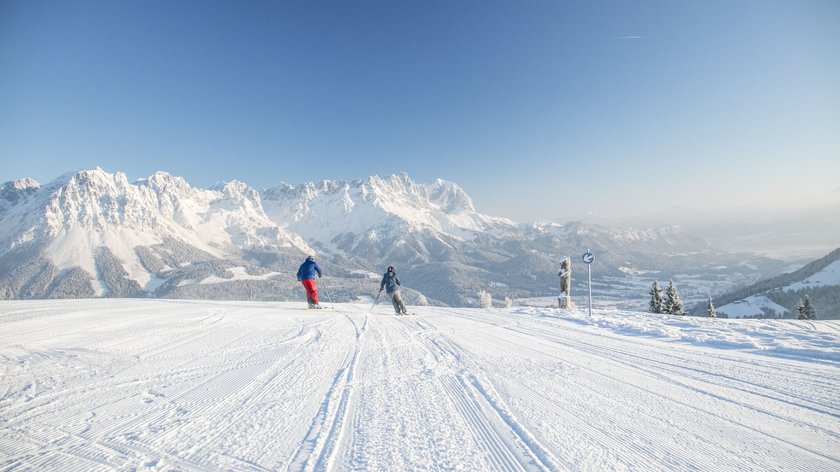 Environmentally friendly skiing fun! Reach our ski region by train Two skiers on snowy slope with Alps in background under clear blue sky