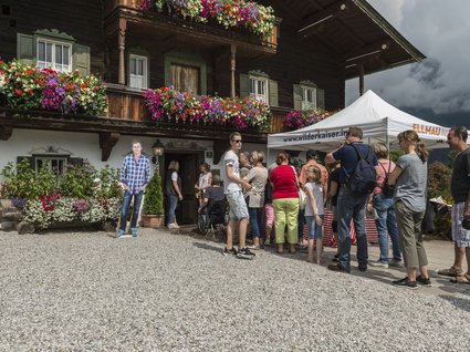 Die Drehorte von “Der Bergdoktor“ am Wilden Kaiser Menschen stehen in einer Schlange vor einem Zelt und einem traditionellem Holzhaus mit Blumen