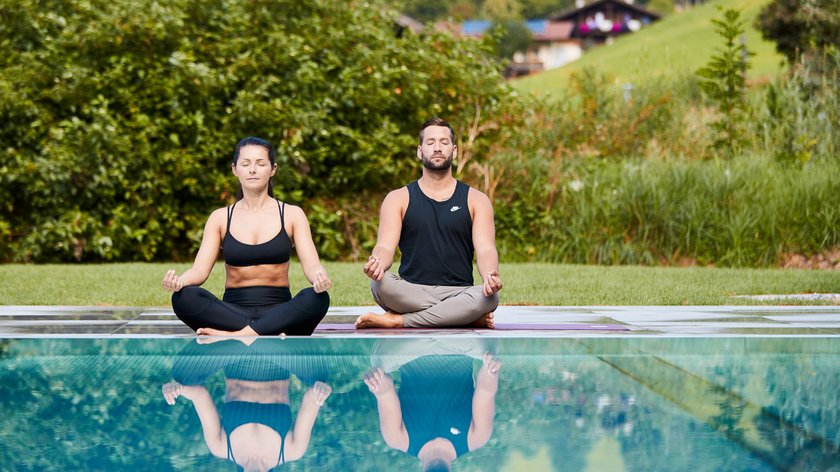 Lohnt sich ein Yoga-Urlaub? Paar meditiert im Lotussitz vor einem Pool in grüner Landschaft