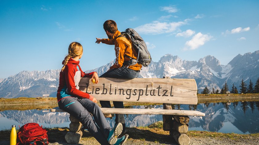 Herbstliche Veranstaltungen in Söll Zwei Wanderer auf Bank vor Bergsee und Alpen mit Aufschrift Lieblingsplatzl