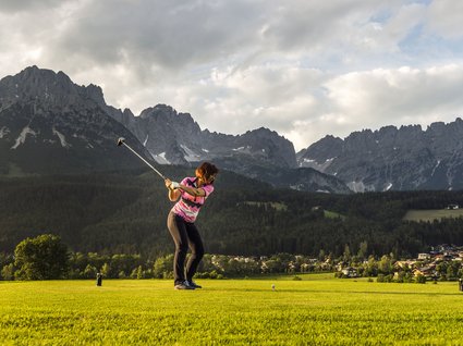 Discover the benefits of the Wilder Kaiser GästeCard to be used in your holiday area- Woman playing golf with mountainous landscape and cloudy sky in background