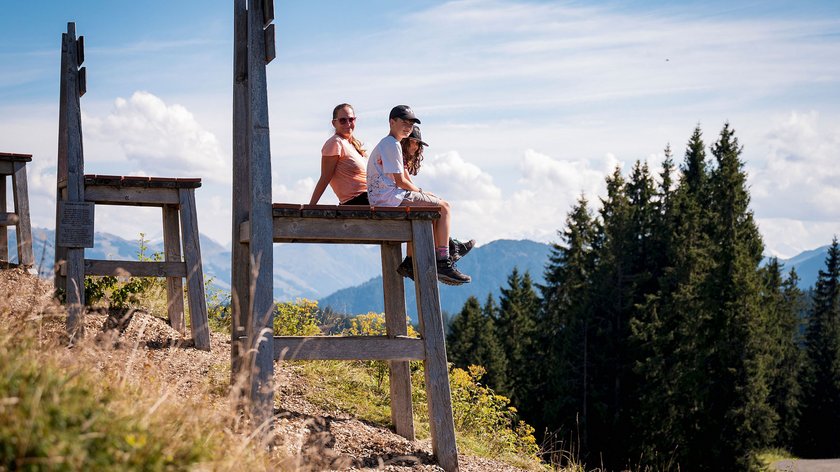 Abenteuerspaß für die ganze Familie Drei Personen sitzen auf übergroßem Stuhl mit Bergblick bei Sonnenschein.