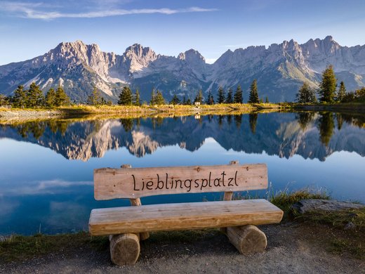 Discover the benefits of the Wilder Kaiser GästeCard to be used in your holiday area- Bench with Lieblingsplatz sign in front of mountain lake reflecting jagged peaks