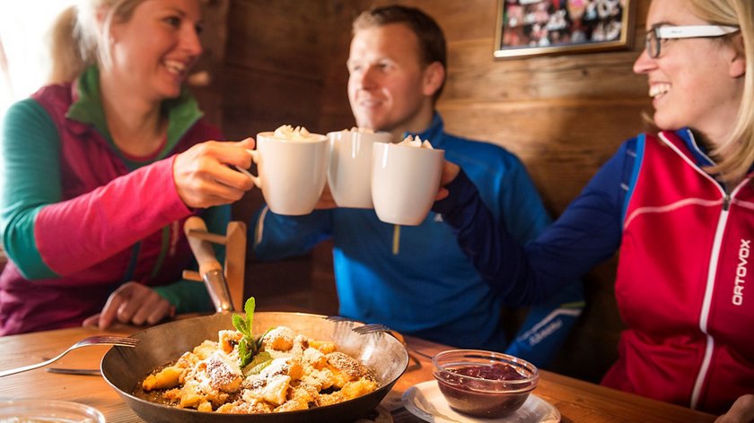 Winterabenteuer mit der ganzen Familie Drei Freunde stoßen mit heißen Getränken an, Person vor Pfannengericht mit Soße