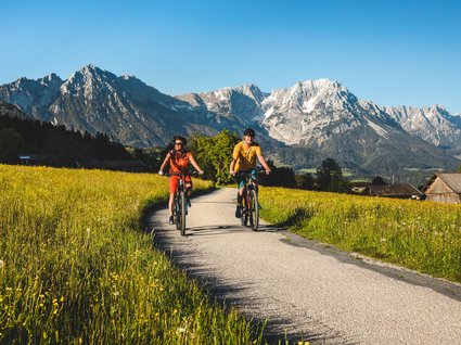 Discover the benefits of the Wilder Kaiser GästeCard to be used in your holiday area- Two cyclists on country road with mountain backdrop and green meadows with flowers