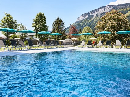 Holidays at the Postwirt: more than just in “a” hotel in Soll. Outdoor pool with lounge chairs and umbrellas with mountains in background on sunny day