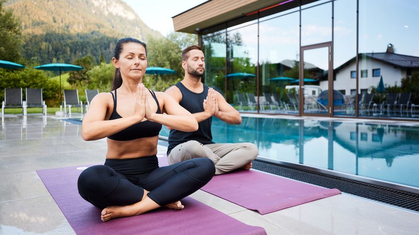 Was ist Hatha Yoga? Paar meditiert im Lotussitz auf Yogamatten neben einem Pool mit Bergblick