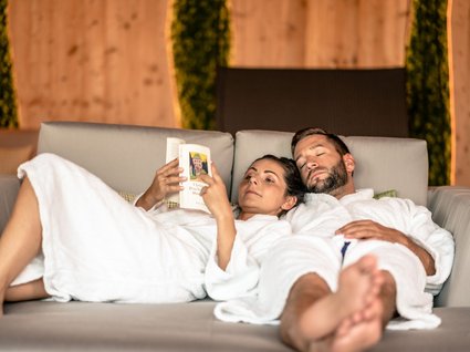 Entspannung im Wellnessbereich für Paare Couple in bathrobes relaxing on sofa, woman reading book, man sleeping