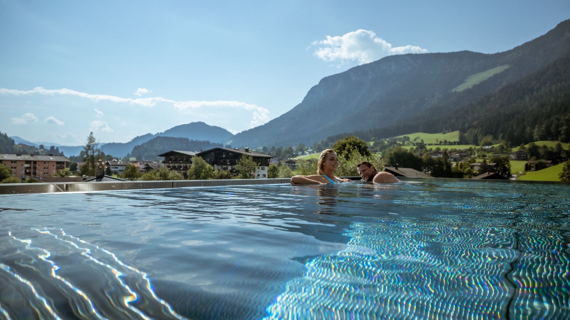 Holidays at the Postwirt: more than just in “a” hotel in Soll. Couple relaxing in infinity pool with mountain view on a sunny day