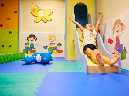 Kind rutscht im Indoor-Spielbereich Ein Kind rutscht glücklich auf einer kleinen Rutsche in einem bunt gestalteten Indoor-Spielbereich mit Kletterwand und weichen Matten.