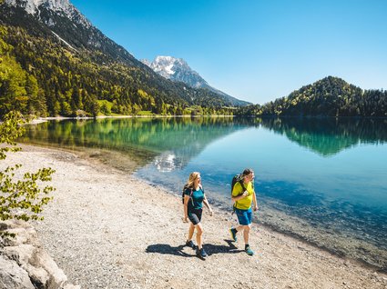 Discover the benefits of the Wilder Kaiser GästeCard to be used in your holiday area- Couple hiking along a mountain lake shore with clear water and sunny sky