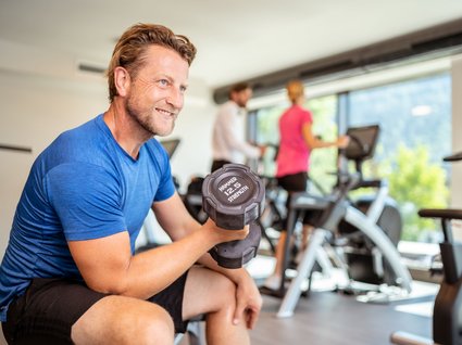Sie suchen ein Hotel mit Fitnessstudio in Österreich? Mann hebt 12,5 kg Hantel im Fitnesstudio mit anderen Trainierenden im Hintergrund